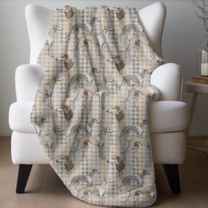 May include: A soft, beige and gray throw blanket with a houndstooth pattern and a repeating image of greyhounds wearing brown and white sweaters. The blanket is draped over a white armchair.