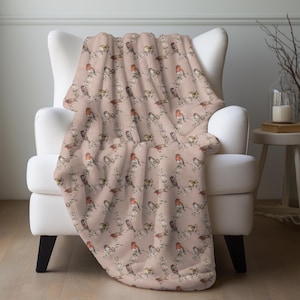 May include: A soft, patterned throw blanket draped over a white armchair. The blanket features a repeating design of colorful birds perched on branches against a pale pink background. The birds have red, brown, and yellow feathers.