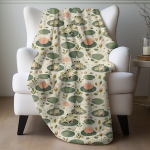 May include: A soft, cream-colored throw blanket with a repeating pattern of green frogs sitting on lily pads with pink flowers. The blanket is draped over a white armchair.