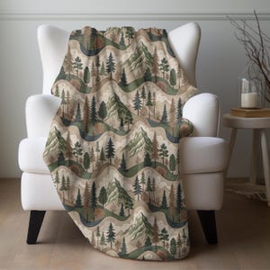 May include: A soft, brown and green throw blanket with a repeating pattern of stylized mountains, trees, and clouds. The blanket is draped over a white armchair.