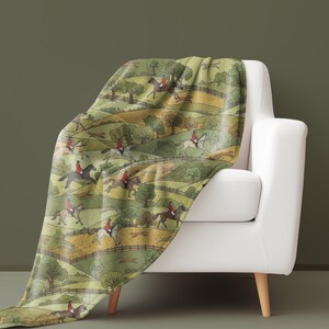 May include: A green and yellow throw blanket draped over a white armchair. The blanket features a repeating pattern of riders on horseback, trees, and rolling hills. The riders wear red jackets.