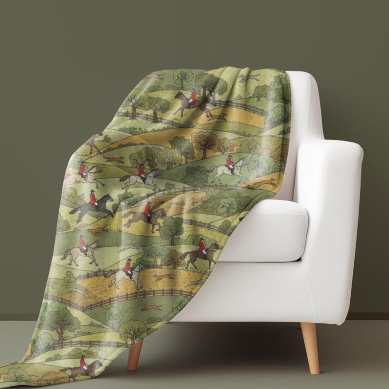 May include: A green and yellow throw blanket draped over a white armchair. The blanket features a repeating pattern of riders on horseback, trees, and rolling hills. The riders wear red jackets.