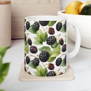 May include: White ceramic mug with a black and green pattern of blackberries. The mug has a handle on the right side.