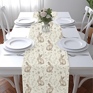 May include: A white table runner with a beige floral pattern featuring bunnies and leaves. The runner is on a white table with two white plates and forks on each side.