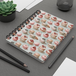 Op de afbeelding: Een spiraalgebonden notitieboek met een lichtblauwe omslag met een herhalend patroon van aquarel macarons met roze, witte en blauwe glazuur en bessen.