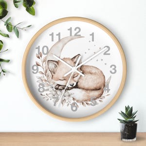 May include: A round wall clock with a light wood frame. The clock face features a sleeping fox curled up on a crescent moon, surrounded by floral accents. The numbers are gray, and the hands are black.