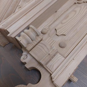 Op de afbeelding: Close-up van onafgewerkte houten architecturale elementen. De stukken hebben ingewikkelde houtsnijwerken, waaronder decoratieve lijsten, afgeronde accenten en gedetailleerde randen. De natuurlijke houtnerf is zichtbaar.