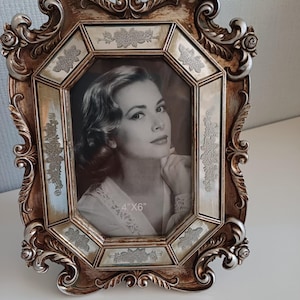 Pode incluir: Moldura de fotos ornamentada em estilo vintage, em tons de bronze e prata. A moldura contém um retrato a preto e branco e mede 10 x 15 cm. A moldura apresenta detalhes florais e de volutas, adicionando um toque de elegância.