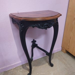 Może przedstawiać: Czarny, drewniany stolik konsolowy z zakrzywionym blatem i ozdobnymi detalami. Stolik ma ciemny drewniany blat i trzy nogi.