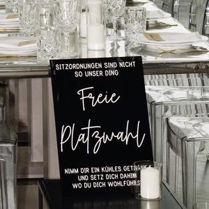 以下が含まれることがあります： 「SITZORDNUNGEN SIND NICHT SO UNSER DING Freie Platzwahl NIMM DIR EIN KÜHLES GETRÄNK UND SETZ DICH DAHIN WO DU DICH WOHLFÜHLS」と書かれた白い文字の黒い看板。白いろうそくがガラスのホルダーに入っています。