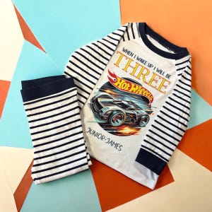 May include: A white and navy blue striped pajama set. The long-sleeved top features a Hot Wheels graphic with the text "WHEN I WAKE UP I WILL BE THREE". The matching pants are navy blue and white striped.