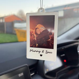 May include: A white, rectangular car air freshener with a photo of two people hugging. The photo is in a polaroid style with the text "Mummy & Isaac" and a heart symbol. The air freshener hangs from a string.