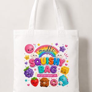 Peut inclure: Un sac fourre-tout blanc avec un motif coloré représentant un arc-en-ciel, des nuages et divers personnages de dessin animé, dont une fraise, du raisin, un beignet et un dinosaure. Le texte indique "maddie SQUISHY BAG FILLED WITH SQUISHY JOY!".