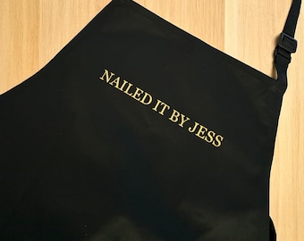 Könnte beinhalten: Schwarze Schürze mit dem goldenen Text "NAILED IT BY JESS". Die Schürze hat einen Nackenriemen und eine verstellbare Schnalle. Die Schürze besteht aus einem glatten, dunklen Material.