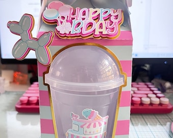 Könnte beinhalten: Eine rosa-weiß gestreifte Pappbox mit einer blauen Schleife oben. Die Box hat ein transparentes Fenster, das einen durchsichtigen Plastikbecher mit einem rosa-blauen Eiswagen-Aufkleber zeigt. Die Box hat außerdem einen goldenen Rand und einen rosa-weißen Ballon in Form eines Hundes. Der Text "Happy Bday" befindet sich auf der Vorderseite der Box.