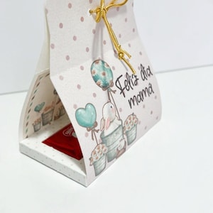 May include: A white gift box with a pink polka dot pattern and a gold ribbon. The box has a cut-out window that reveals a red chocolate bar. The box is decorated with a cartoon bunny holding balloons and flowers. The text "Feliz dia mama" is printed on the box.