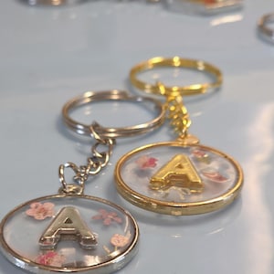 May include: A selection of keyrings featuring the letter "A". Two keyrings are in focus, one with a silver-coloured border and the other gold. Both have a clear resin centre with dried flowers and the letter "A".