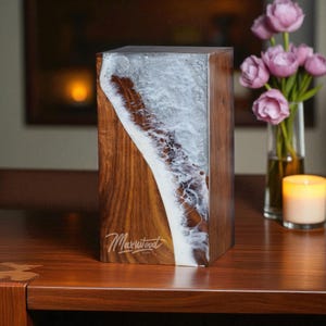 May include: A rectangular wooden block with a unique design. The block features a combination of dark wood and a white, swirling resin pattern, resembling ocean waves. The Maxwood logo is visible on the wood. The block is on a wooden surface.