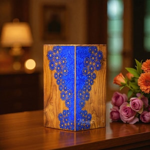 Op de afbeelding: Een rechthoekige houten urn met een verticale inleg van levendig blauwe hars. Het harsgedeelte is versierd met een herhalend patroon van kleine bloemen met donkere randen. De natuurlijke houtnerf is zichtbaar aan de zijkanten.