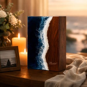 Op de afbeelding: Een rechthoekige houten urn met een donkerbruine houtnerf aan de rechterkant en een blauw hars oceaangolfontwerp met wit schuim aan de linkerkant. De tekst Always in our hearts is in cursief schrift in het houten gedeelte gegraveerd.