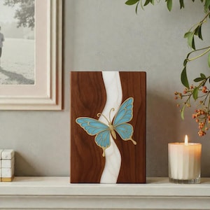 Könnte beinhalten: Rechteckige Holzplakette mit Schmetterlingsdesign. Die Plakette hat einen braunen Holzmaserungshintergrund mit einem weißen, wellenförmigen Streifen in der Mitte. Ein blauer Schmetterling mit goldenen Verzierungen ist auf der Plakette zentriert. Eine brennende Kerze ist im Hintergrund.