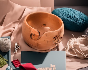 Peut inclure: Un bol à laine en bois avec un motif floral. Le bol est brun clair et a un trou au centre pour enrouler la laine. Le bol est posé sur une surface brun clair avec une pelote de laine turquoise et un écheveau de laine blanche.