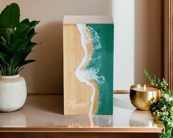 May include: A wooden box with a resin inlay depicting a beach scene with waves crashing on the shore. The box is light brown with a green and white resin inlay. The text "Madewood" is printed on the front of the box.