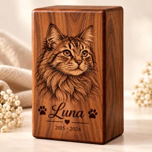 Könnte beinhalten: Eine rechteckige Urne aus Holz mit einem lasergravierten Porträt einer langhaarigen Katze. Unter der Katze ist der Name Luna in Schreibschrift eingraviert, flankiert von zwei Pfotenabdrücken, einem Herz und den Jahren 2015-2024.