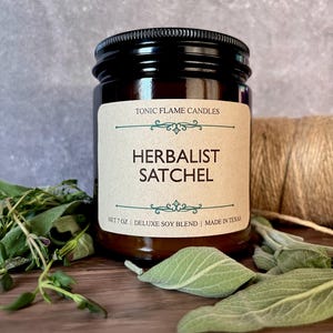 Puede incluir: Un tarro de vidrio marrón con tapa negra, etiquetado "Herbalist Satchel" de Tonic Flame Candles. La etiqueta es beige con texto verde y marrón. La vela está rodeada de hierbas frescas y cuerda. Peso neto 198 gramos.