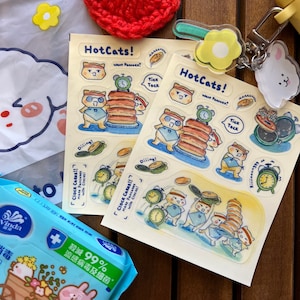 Puede incluir: Dos hojas de pegatinas con el texto "HotCats!" e ilustraciones de personajes de gatos haciendo panqueques. Las pegatinas presentan relojes, panqueques y bocadillos con frases como "Want Pancake?" y "Tick Tock."