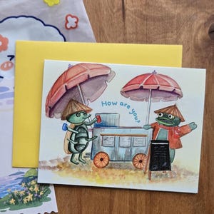 Peut inclure: Carte de vœux aquarelle avec deux créatures anthropomorphes, l'une insectoïde et l'autre en forme de grenouille, sous des parapluies rouges. La carte porte l'inscription "How are you ?" et est accompagnée d'une enveloppe jaune et d'un petit sac.