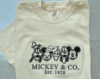 May include: A cream colored t-shirt with a black embroidered image of Mickey Mouse, Donald Duck, Goofy, and Pluto. The text "MICKEY & CO. EST. 1928" is also embroidered in black.