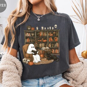 May include: Dark gray t-shirt featuring a Halloween-themed graphic. The design includes a ghost reading in a cozy chair in front of a bookshelf, with pumpkins, skulls, and a black cat. The word "PEPPER" is in a gray circle.