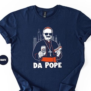 May include: Navy blue t-shirt featuring a cartoon illustration of a figure in religious garb, holding a beer and a hot dog. The text "DA POPE" is printed in white below the image. The Chicago skyline is in the background.