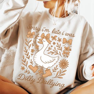May include: Beige sweatshirt with a graphic of a white goose wearing a bow and boots, surrounded by floral elements. The text "Sorry I'm late I was Dilly Dallying" is printed on the front. A black mug is held up.