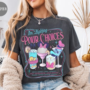 May include: Dark gray t-shirt with the text "I'm Making Pour Choices" and illustrations of cocktails. The shirt has a vintage, worn look and lists several countries. A woman is wearing the shirt.