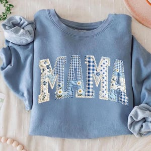 May include: A light blue sweatshirt with the word "MAMA" appliqued across the front. The letters are a patchwork of floral, gingham, and polka dot patterns in shades of blue and white. The sleeves are rolled up.