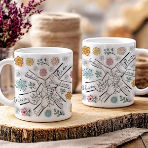 May include: Two white ceramic mugs with black outlines of hands holding each other. The mugs are decorated with colorful flowers and the names "Olivia", "Sarah", "Alex", "Liam", "Nana", "Isabella" are written on the mugs.