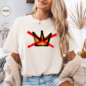 May include: Ivory-colored t-shirt featuring a graphic crown design in black, yellow, and red, crossed out with red lines. The shirt is a relaxed fit. The word "IVORY" is in a circle in the upper left corner.