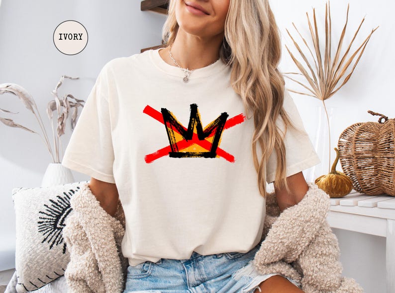 May include: Ivory-colored t-shirt featuring a graphic crown design in black, yellow, and red, crossed out with red lines. The shirt is a relaxed fit. The word "IVORY" is in a circle in the upper left corner.