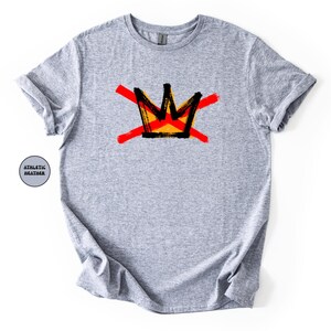 May include: A light gray t-shirt featuring a graphic of a crown design in black, yellow, and red. The crown is crossed out with two red brushstrokes. The shirt is made of athletic heather material.