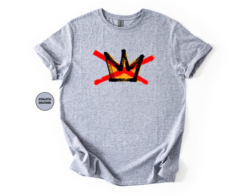 May include: A light gray t-shirt featuring a graphic of a crown design in black, yellow, and red. The crown is crossed out with two red brushstrokes. The shirt is made of athletic heather material.