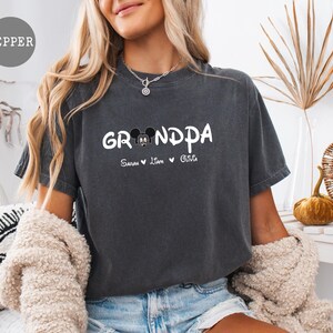 Op de afbeelding: Een donkergrijs T-shirt met het woord "Grandpa" in witte letters en een Mickey Mouse-kopgrafiek. Het shirt heeft ook de namen "Sarah", "Liam" en "Olivia" gedrukt onder de grafiek.