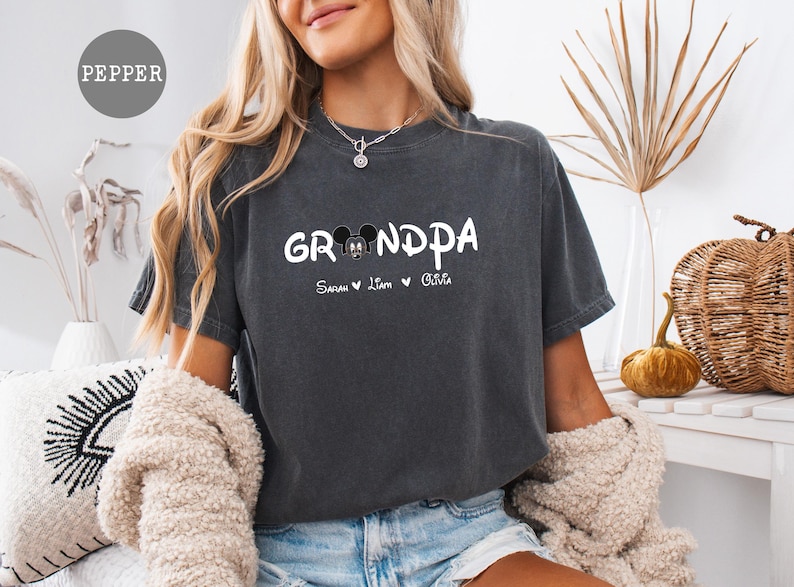 Op de afbeelding: Een donkergrijs T-shirt met het woord "Grandpa" in witte letters en een Mickey Mouse-kopgrafiek. Het shirt heeft ook de namen "Sarah", "Liam" en "Olivia" gedrukt onder de grafiek.