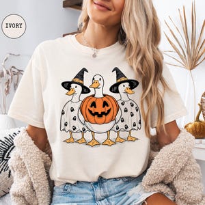 Peut inclure: T-shirt ivoire avec un motif d'Halloween. Le motif repr&eacute;sente trois canards, deux portant des chapeaux de sorci&egrave;re et des costumes de fant&ocirc;mes, et un tenant une citrouille sculpt&eacute;e. Le t-shirt est de coupe d&eacute;contract&eacute;e.