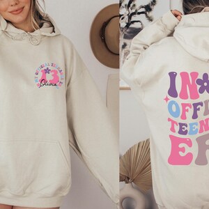 May include: A light beige hoodie with a graphic print on the front and back. The front graphic says "My Official Teenage Era" with a pink number 13 and the name "Olivia". The back graphic says "In My Official Teenager Era" in a colorful retro font.