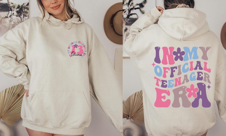 May include: A light beige hoodie with a graphic print on the front and back. The front graphic says "My Official Teenage Era" with a pink number 13 and the name "Olivia". The back graphic says "In My Official Teenager Era" in a colorful retro font.