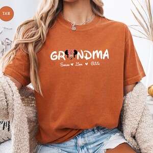 Op de afbeelding: Oranje T-shirt met het woord "Grandma" in witte tekst met een cartoon Minnie Mouse-hoofd in het midden van het woord. De tekst "Sarah Liam Olivia" staat onder het woord "Grandma".