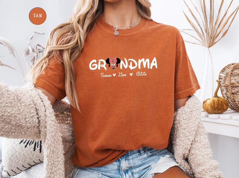 Op de afbeelding: Oranje T-shirt met het woord "Grandma" in witte tekst met een cartoon Minnie Mouse-hoofd in het midden van het woord. De tekst "Sarah Liam Olivia" staat onder het woord "Grandma".