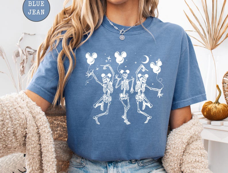 May include: Blue jean colored t-shirt featuring a white graphic of three dancing skeletons holding Mickey Mouse-shaped balloons. The design includes stars and a crescent moon. The shirt has short sleeves and a crew neck.